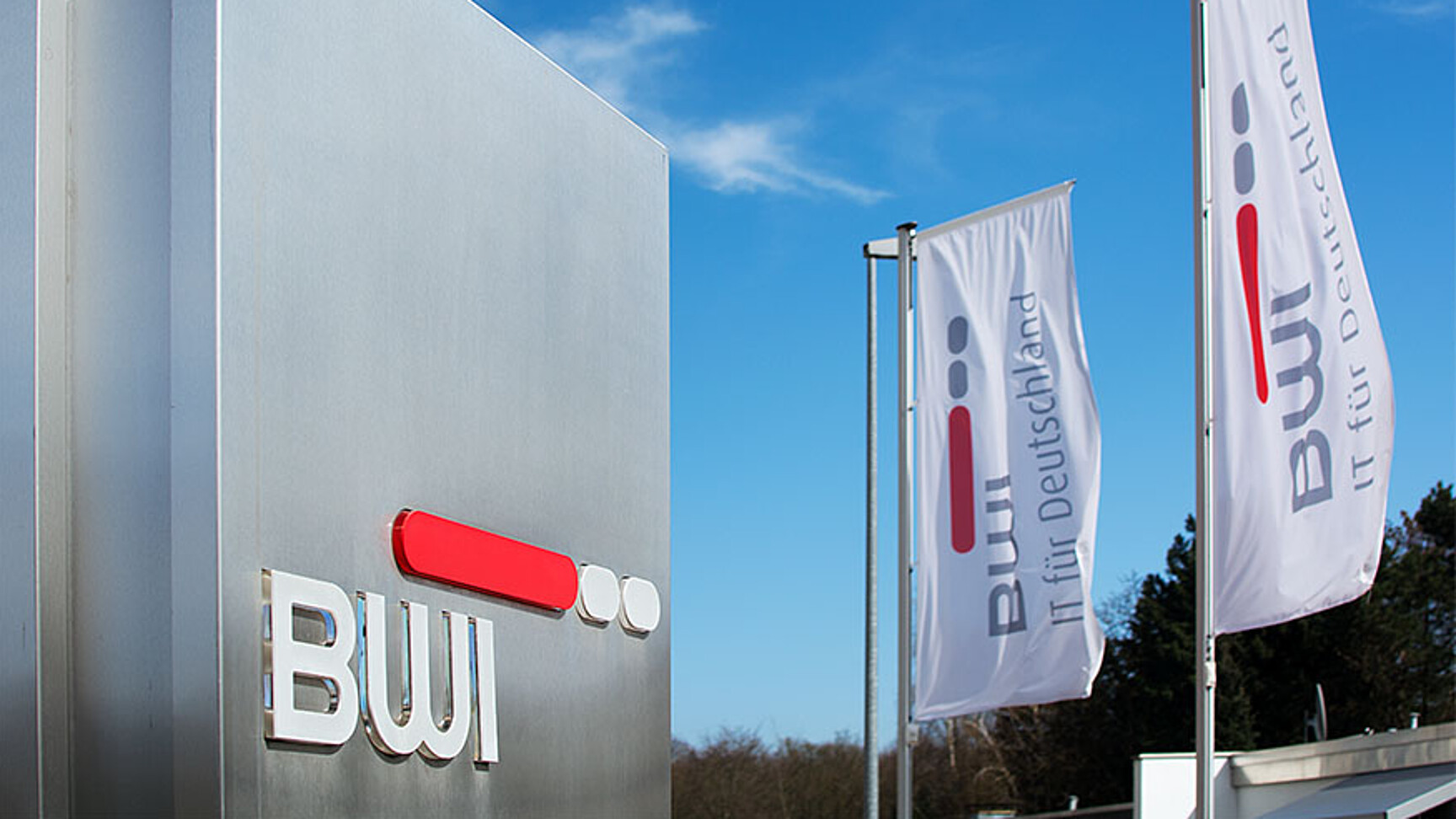 Im Hintergrund zwei weiße Fahnen am Fahnenmast mit BWI Logo. Im Vordergrund rechts eine Stahl Stehle mit BWI Logo. Eingang zu Liegenschaft fotografiert.
