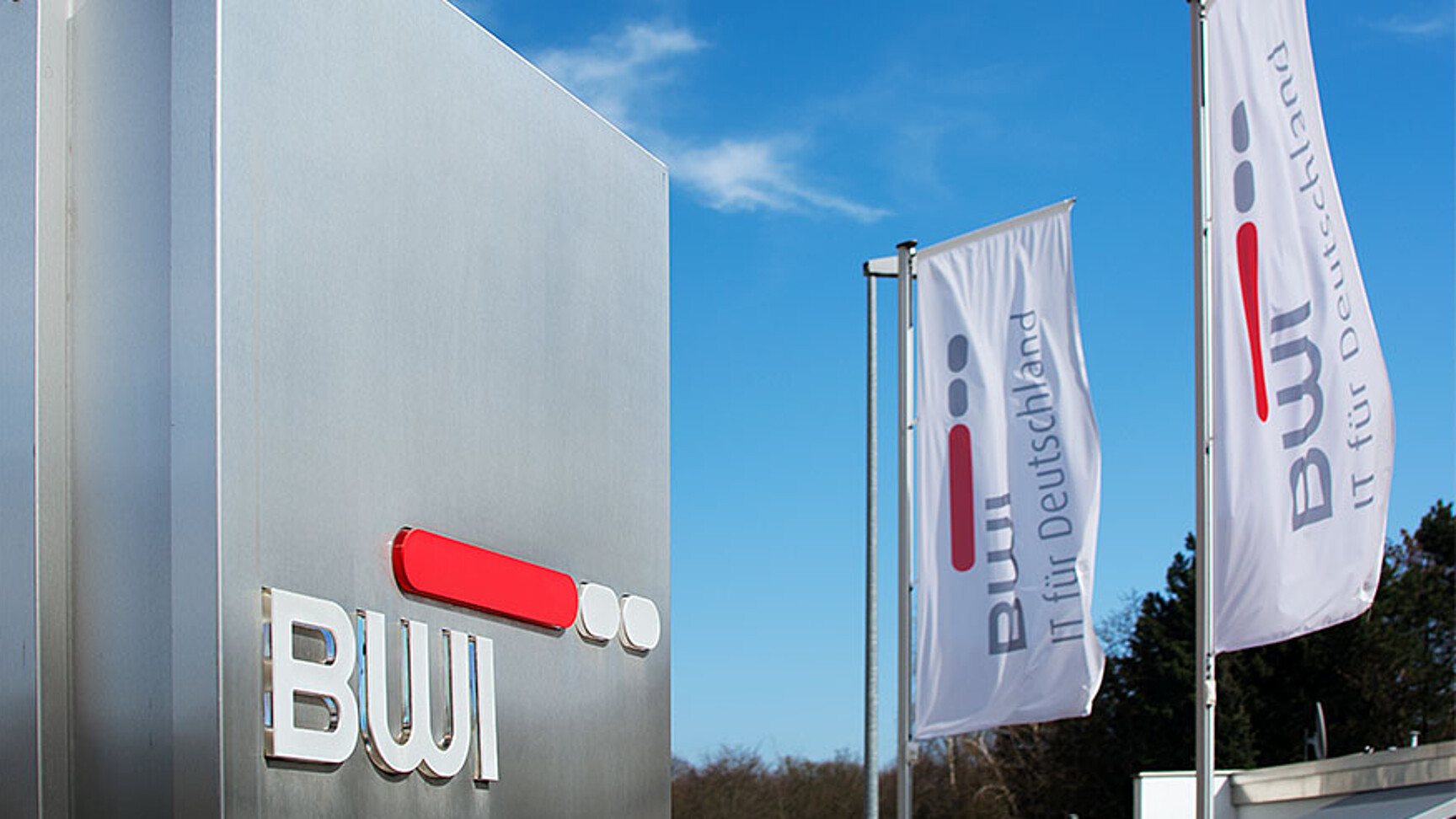 Im Hintergrund zwei weiße Fahnen am Fahnenmast mit BWI Logo. Im Vordergrund rechts eine Stahl Stehle mit BWI Logo. Eingang zu Liegenschaft fotografiert.