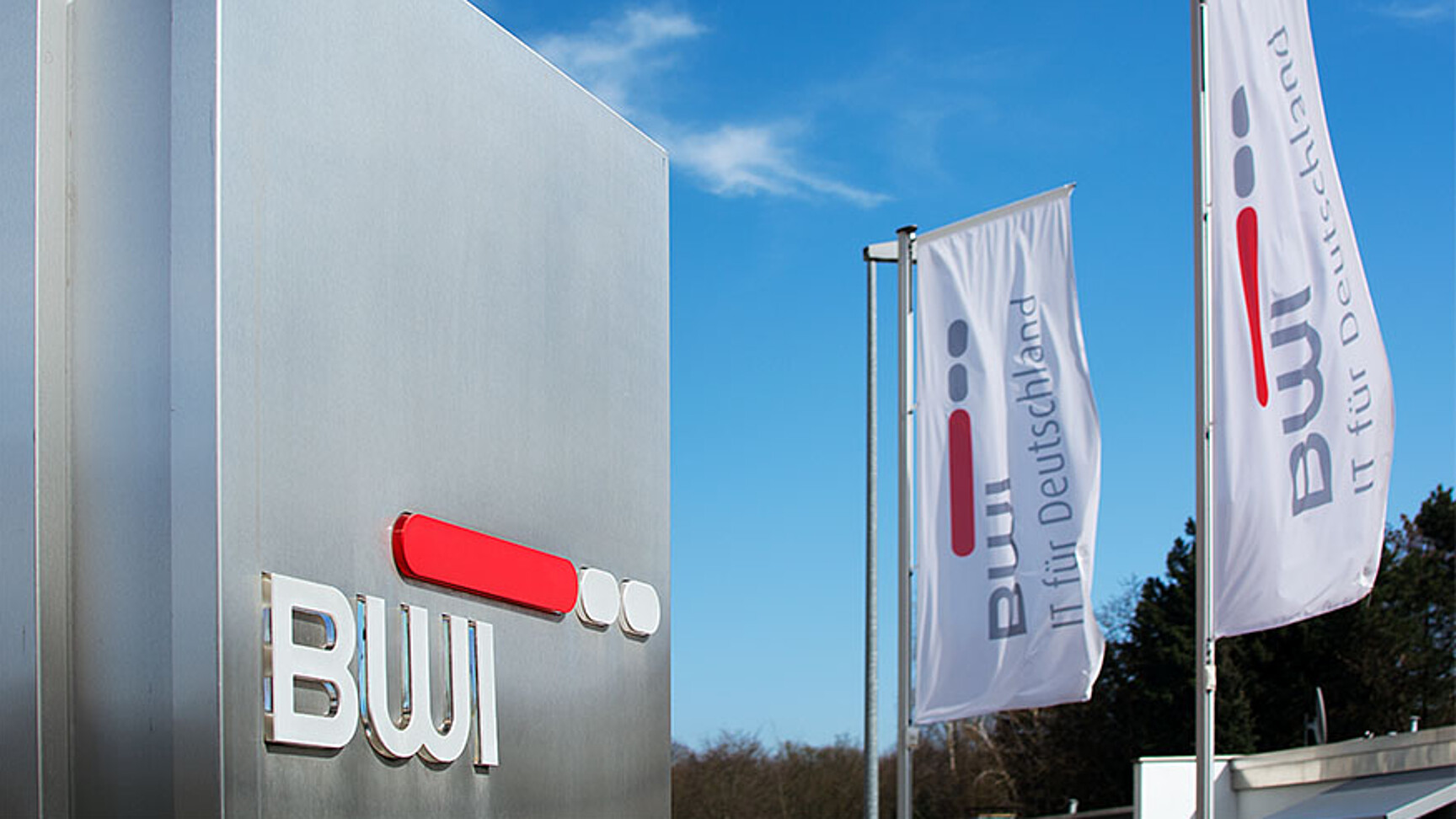 Im Hintergrund zwei weiße Fahnen am Fahnenmast mit BWI Logo. Im Vordergrund rechts eine Stahl Stehle mit BWI Logo. Eingang zu Liegenschaft fotografiert.