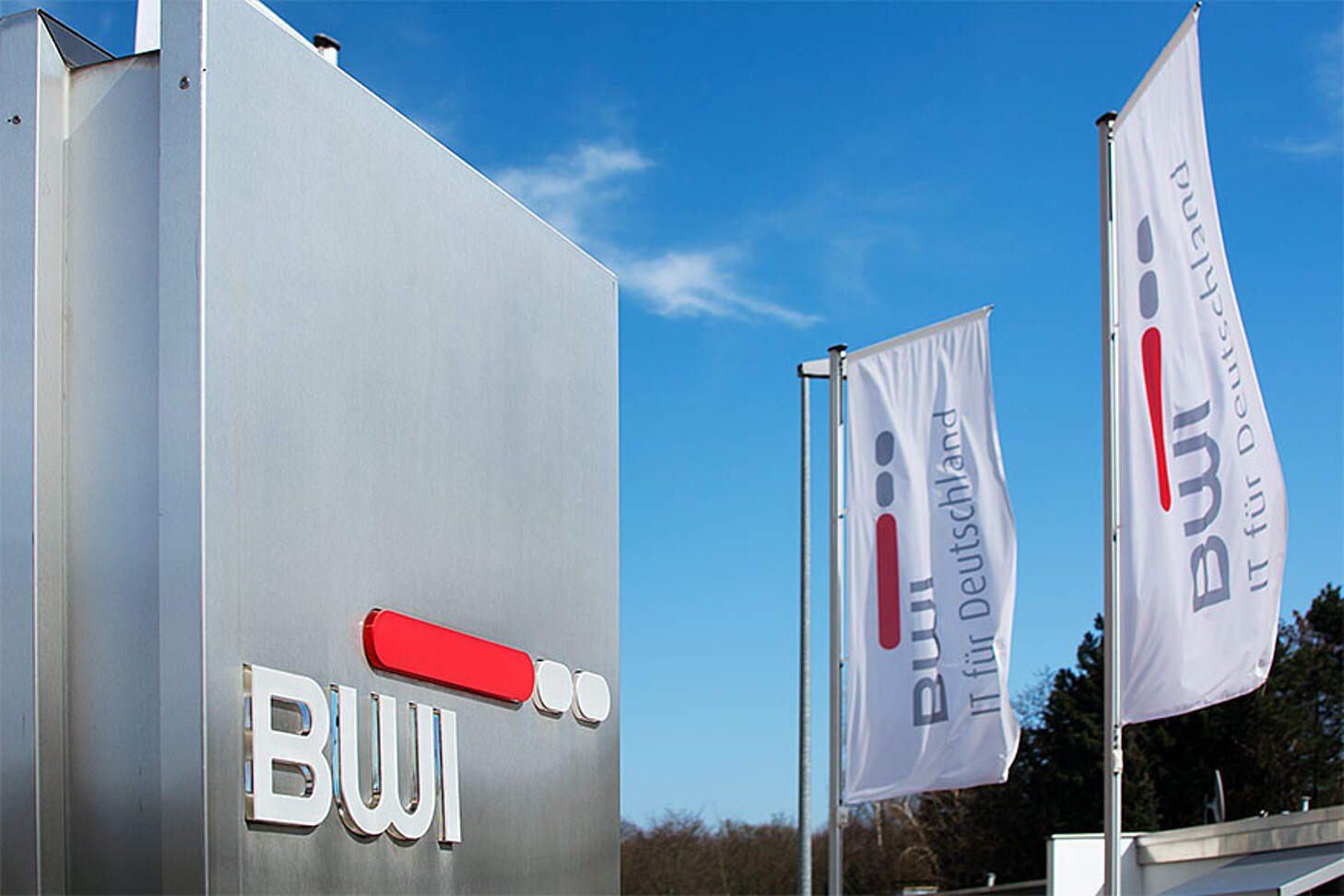 Im Hintergrund zwei weiße Fahnen am Fahnenmast mit BWI Logo. Im Vordergrund rechts eine Stahl Stehle mit BWI Logo. Eingang zu Liegenschaft fotografiert.