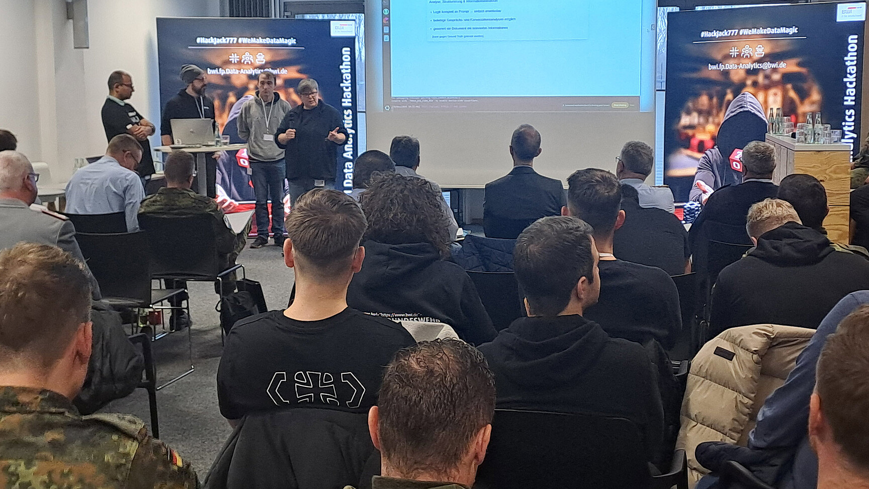 Hackthon Teilnhemer sitzen auf Stühlen und schauen einer Präsentation eines Teams von 4 Leuten zu