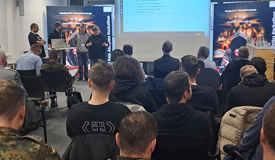 Hackthon Teilnhemer sitzen auf Stühlen und schauen einer Präsentation eines Teams von 4 Leuten zu