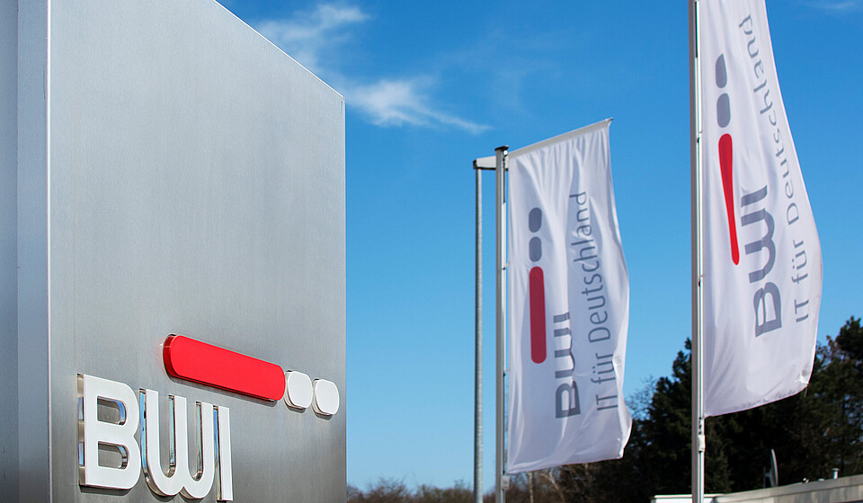 Stele mit BWI-Logo, im Hintergrund zwei gehisste BWI-Fahnen