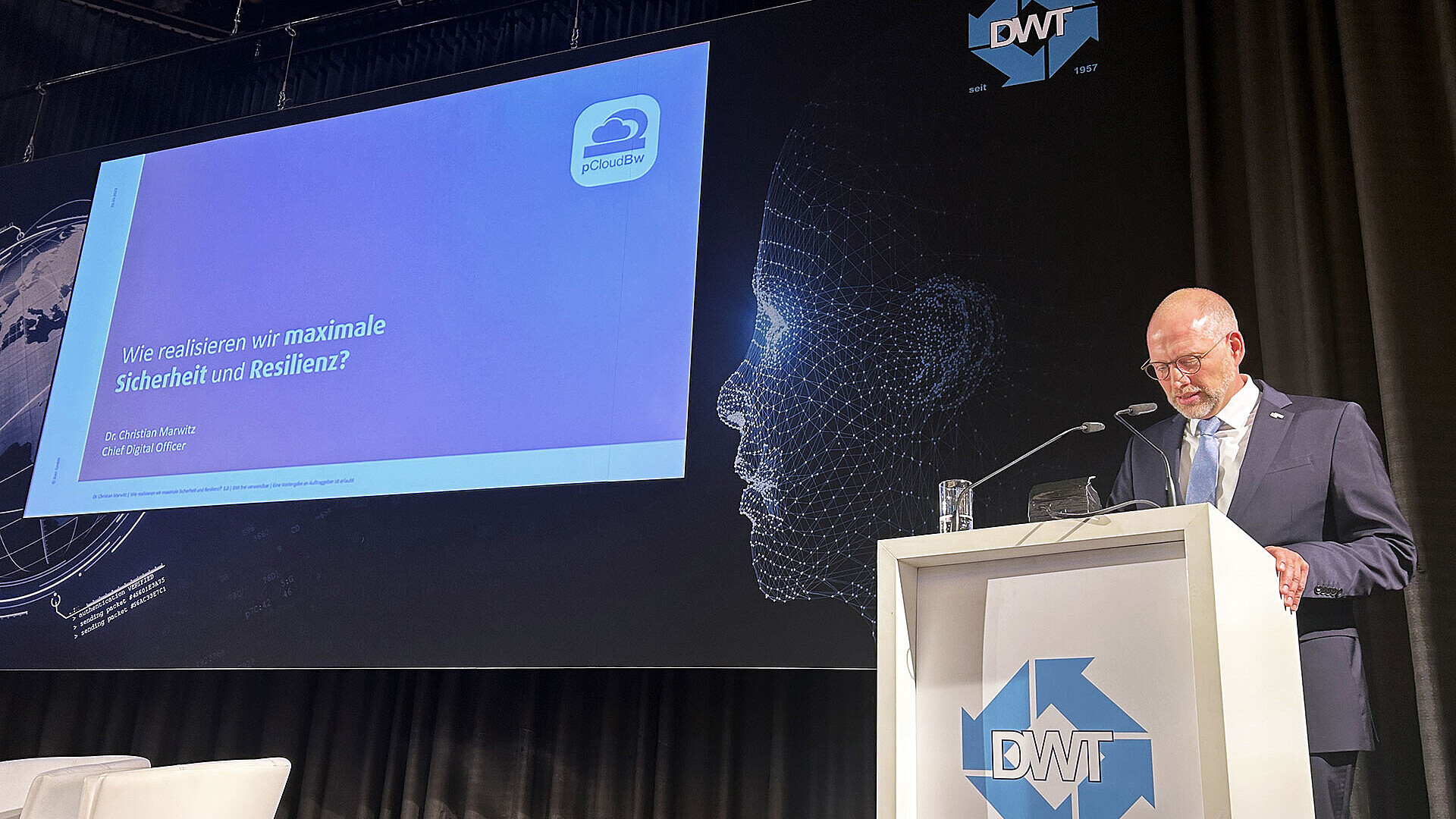 Dr. Christian Marwitz, CDO der BWI, steht auf der Bühne hinter einem Rednerpult während seines Vortrags auf der Cyber Defence Conference 2023; im Hintergrund ist sein Vortragstitel an eine Bühnenrückwand projeziert;