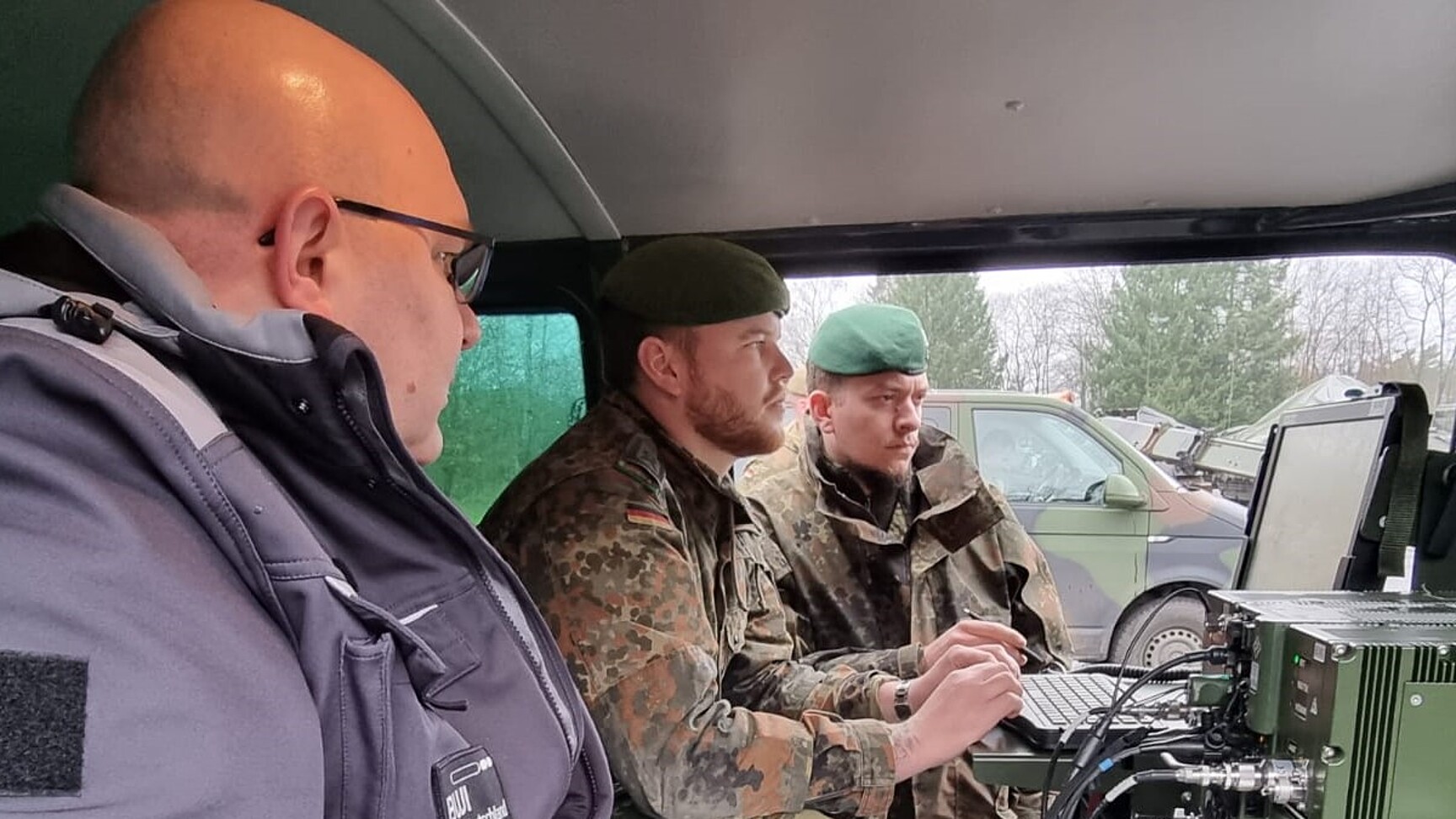 eine BWI Mitarbeiter und zwei Soldaten in Uniform schauen in einem Fahrzeug auf einen großen Monitor.