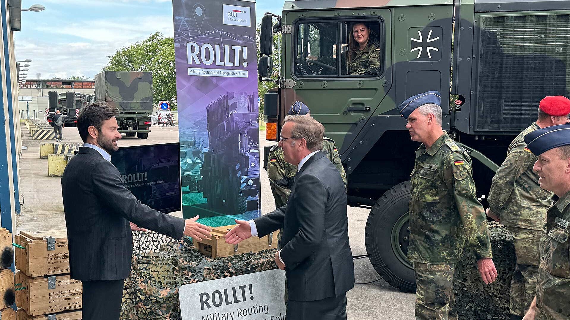 Verteidigungsminister Boris Pistorius schüttelt Gregro Fiedler (Innovationmanager BWI) die Hand. Hinter dem Verteidigungsminister steht Generalmajor Gerald Funke, Kommandeur Logistikkommando der Bundeswehr. Im Hintergrund sind Transportfahrzeuge der Bundeswehr zu sehen.