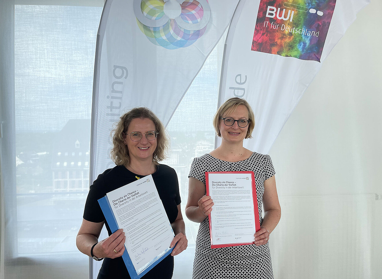 Sandra Bußmann und Katrin Hahn nebeneinander und zeigen die unterzeichnete Charta der Vielfalt ins Bild. Im Hintergrund ist das BWI Logo in den Pride Farben zusehen.