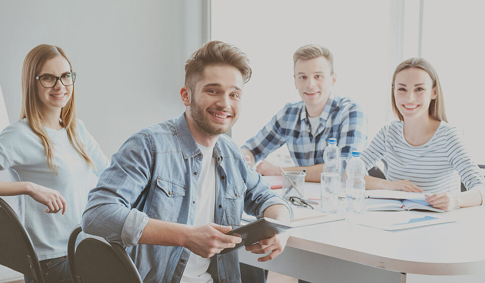 4 junge Personen sitzen an einen Meetingtisch. Alle schauen und lächeln in die Kamera für eine positive Stimmung.