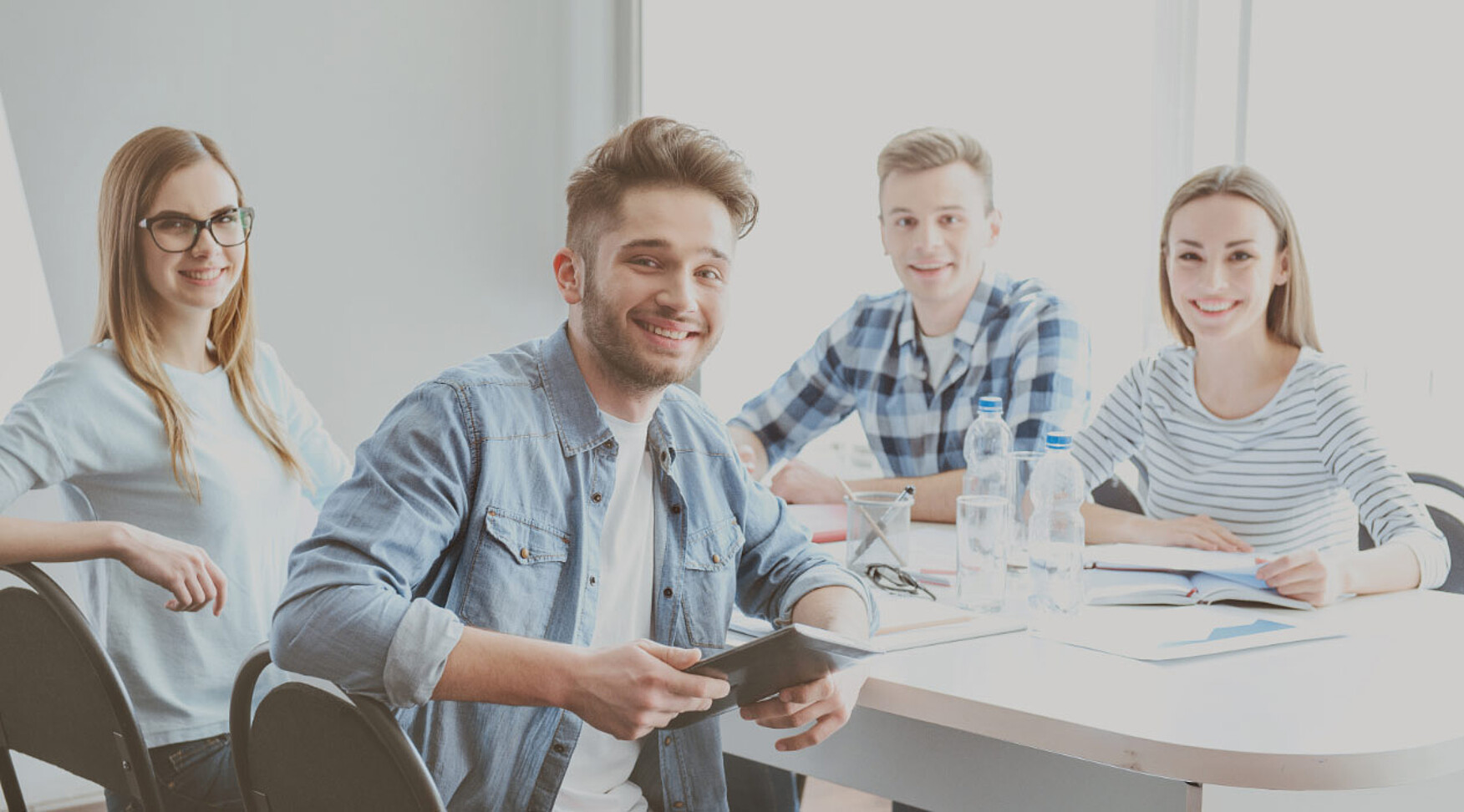 4 junge Personen sitzen an einen Meetingtisch. Alle schauen und lächeln in die Kamera für eine positive Stimmung.